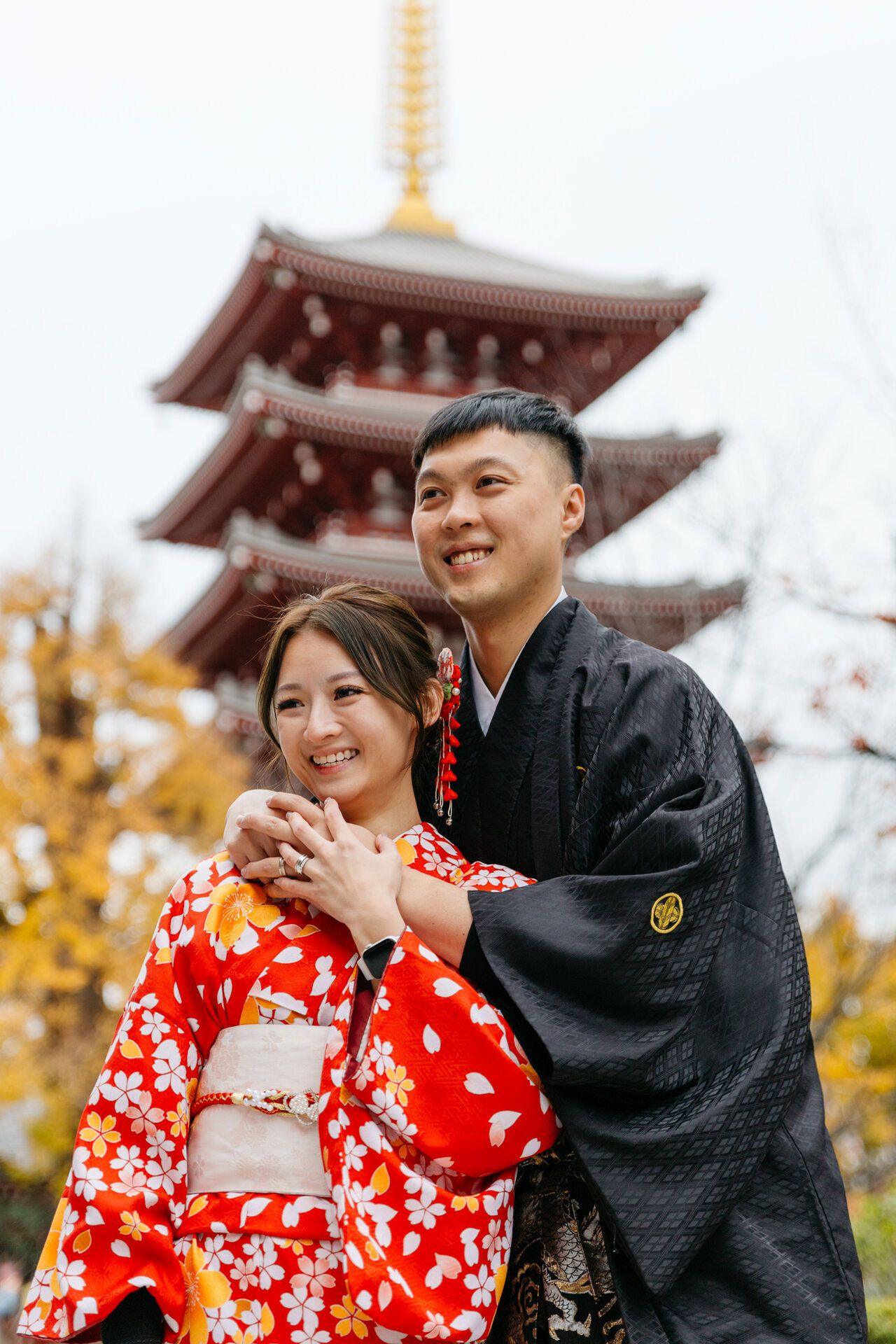 Two people in traditional Japanese attire pose together in front of a pagoda during autumn.