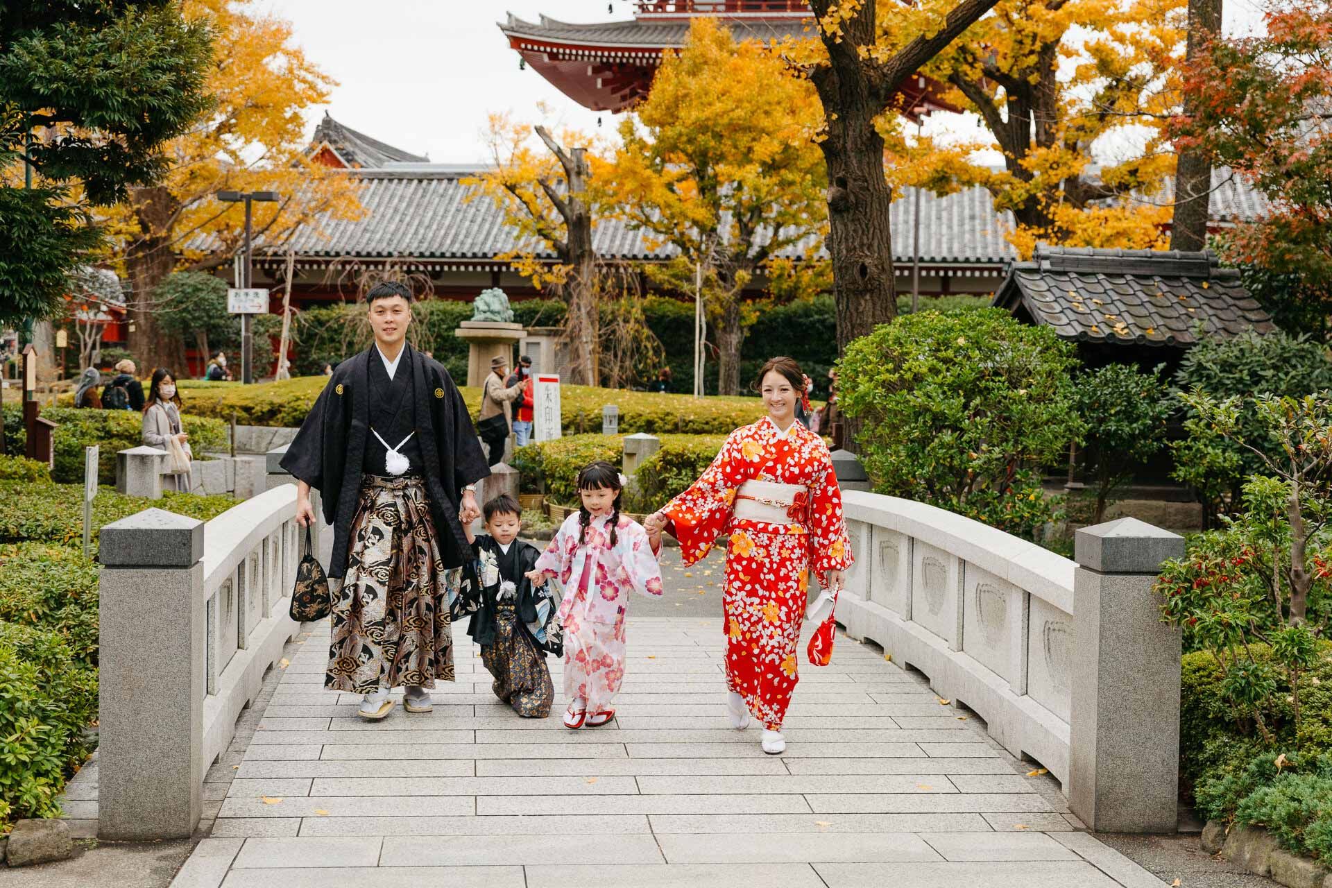 Family walking across a stone bridge in traditional Japanese attire with autumn foliage and temple in background.