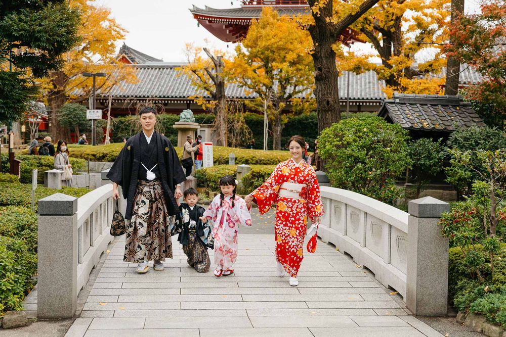 Family walking across a stone bridge in traditional Japanese attire with autumn foliage and temple in background.