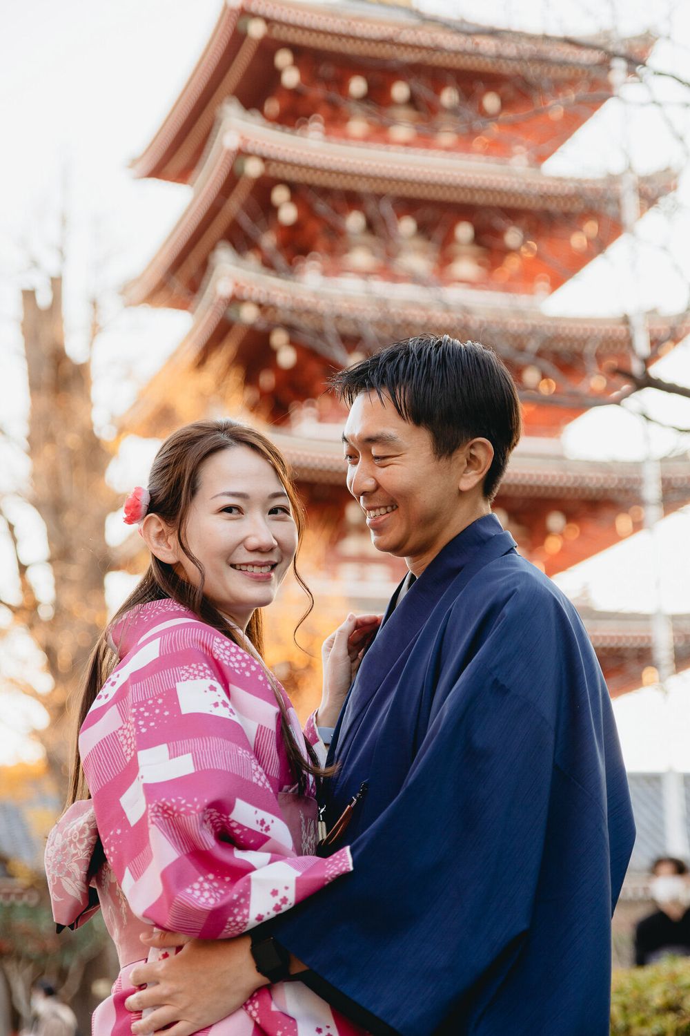 Two people in traditional Japanese dress share a moment in front of a pagoda during autumn.