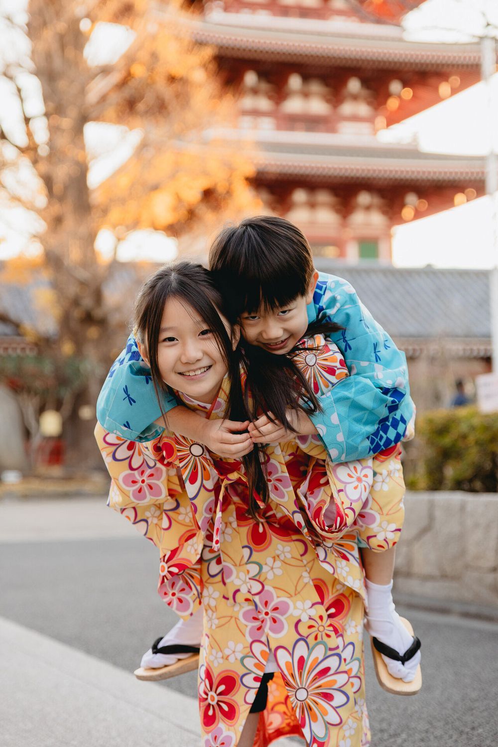 Two people in colorful kimonos share a playful piggyback ride in front of a traditional Japanese temple.