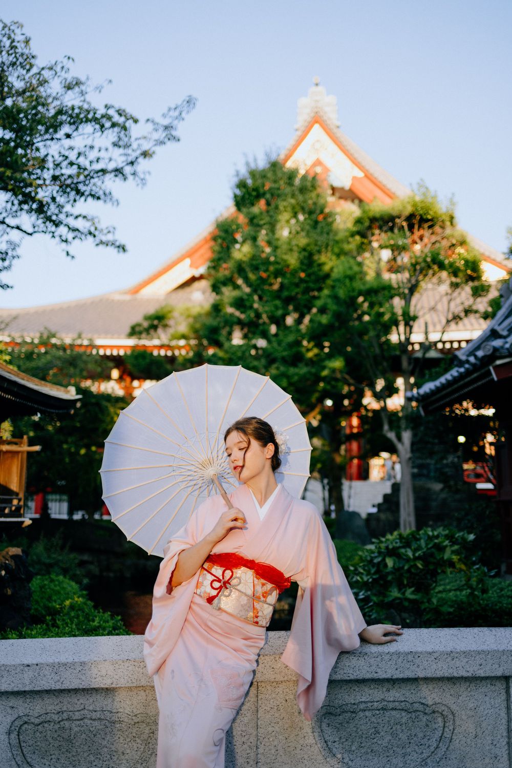 Figure in traditional kimono with white parasol poses in front of Japanese temple architecture at sunset.