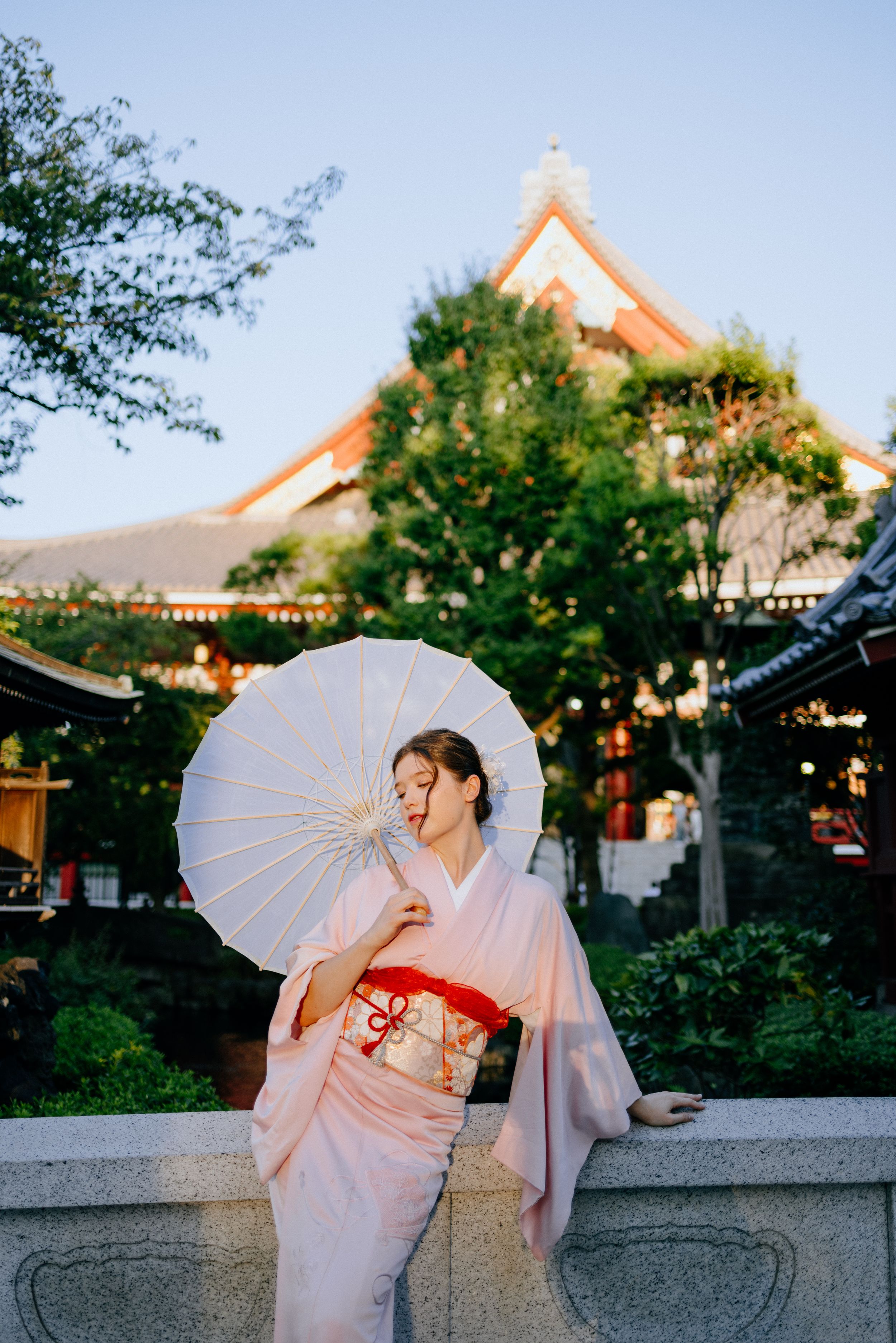 Figure in traditional kimono with white parasol poses in front of Japanese temple architecture at sunset.