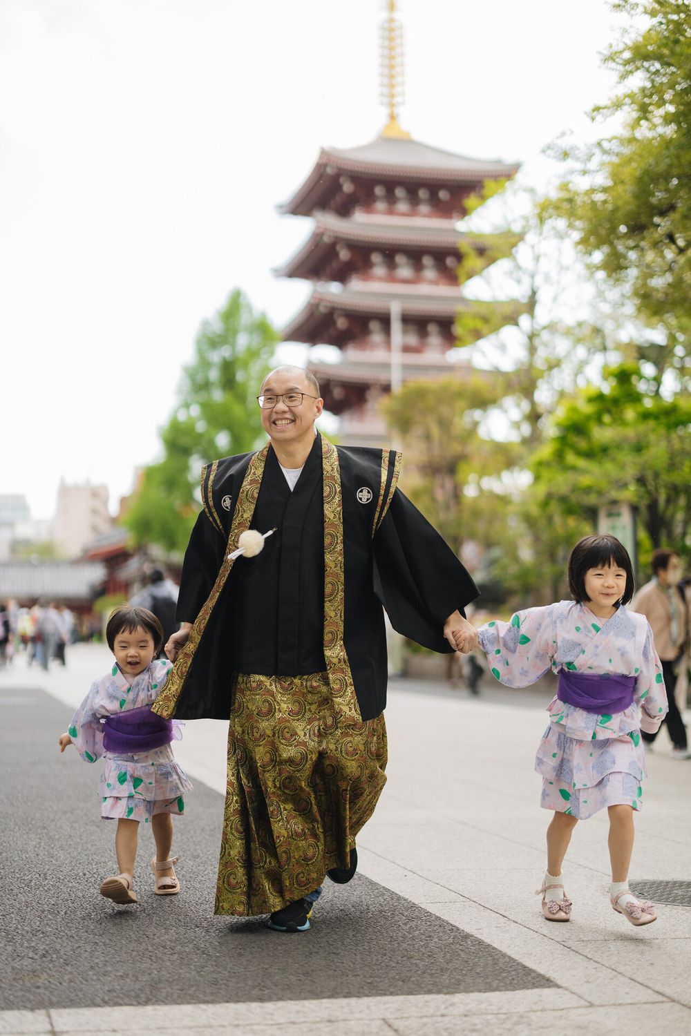 Person in black and gold ceremonial robes walking with children in yukata near pagoda.