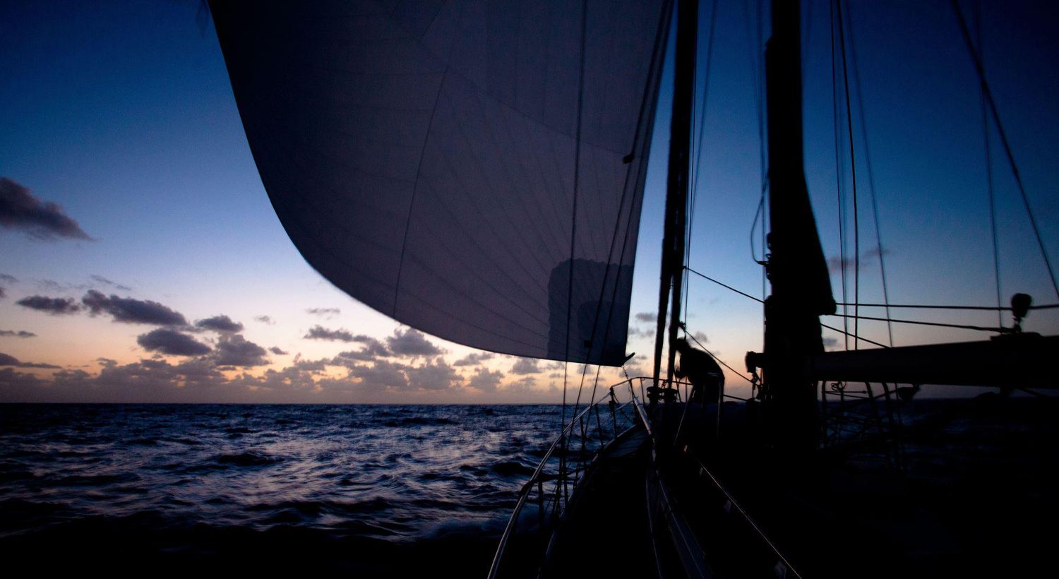 Sunset view from sailboat deck with dark sails silhouetted against twilight sky.