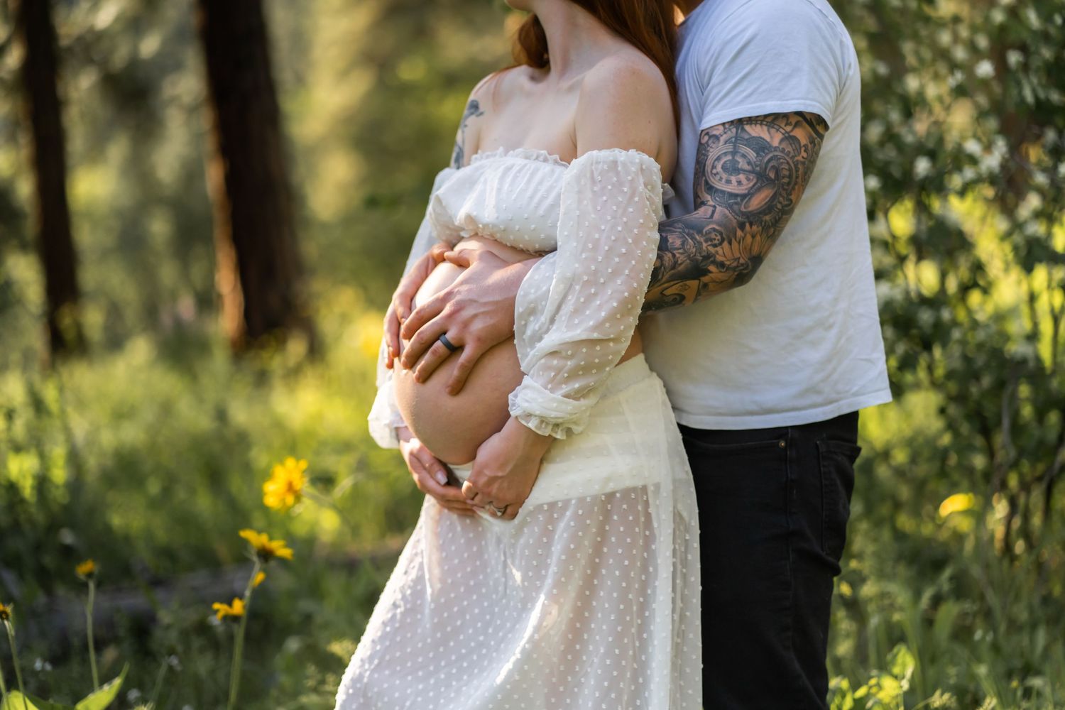 A tender maternity moment captured in a white lace dress against a forest backdrop.