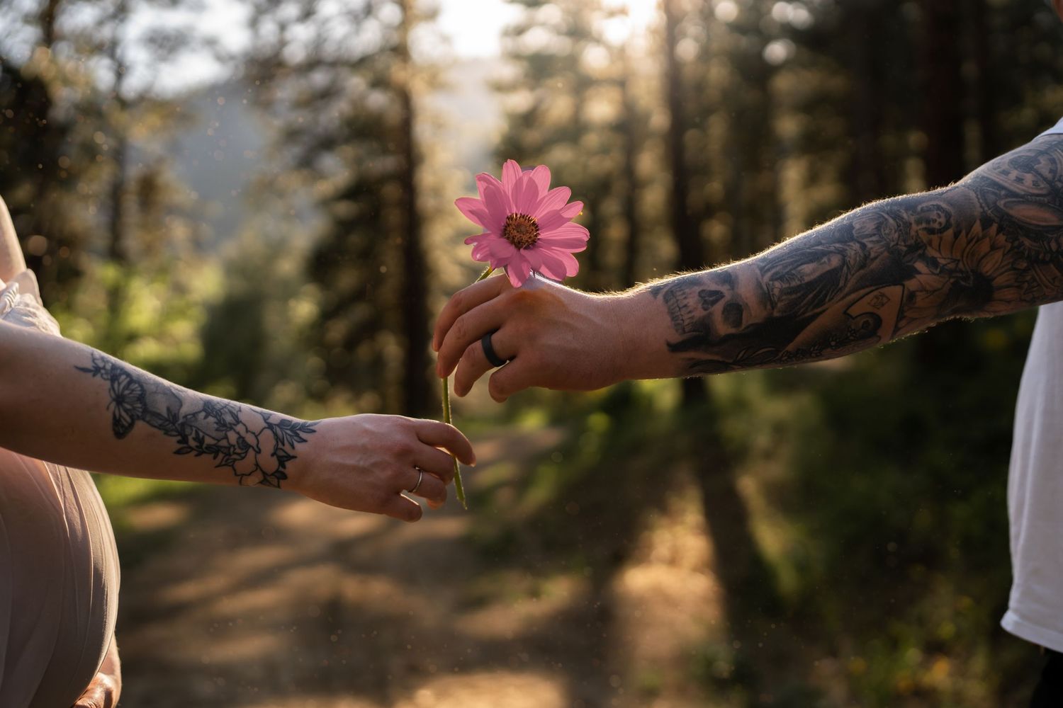Tattooed arms exchange a pink flower in a sunlit forest setting.