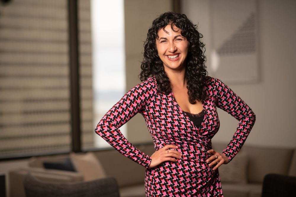 A smiling person poses in a fitted red and white polka dot wrap dress with hands on hips in an office setting.