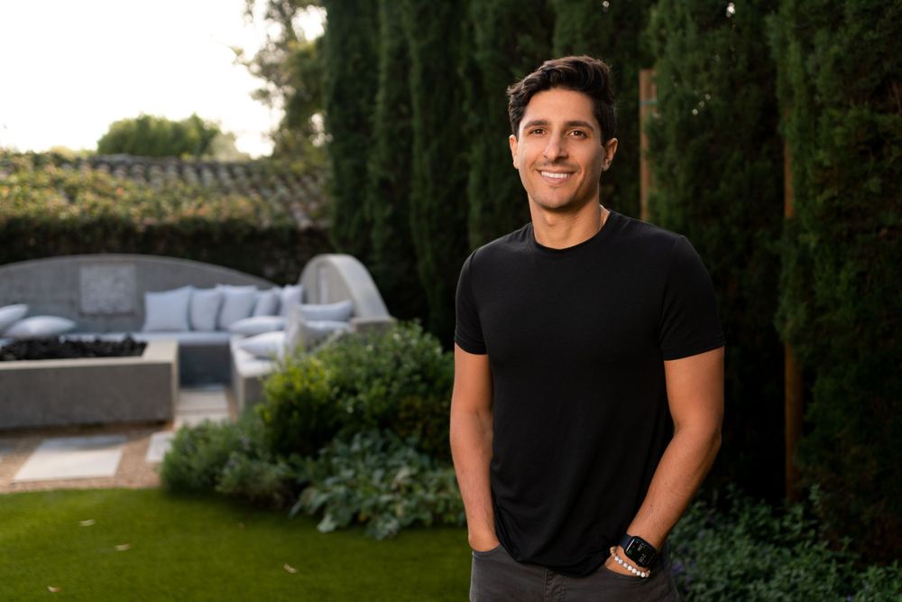 A person in a black t-shirt stands in a manicured garden with cypress trees and stone features at sunset.