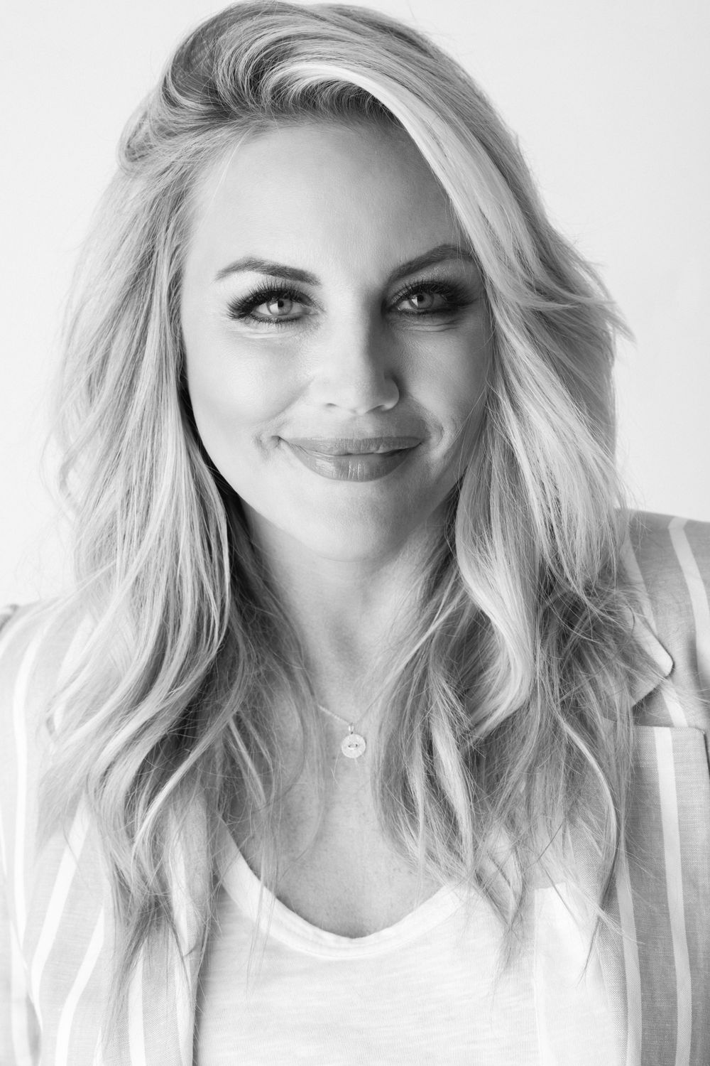 Black and white studio portrait of a person with wavy blonde hair and a bright smile wearing a white top.