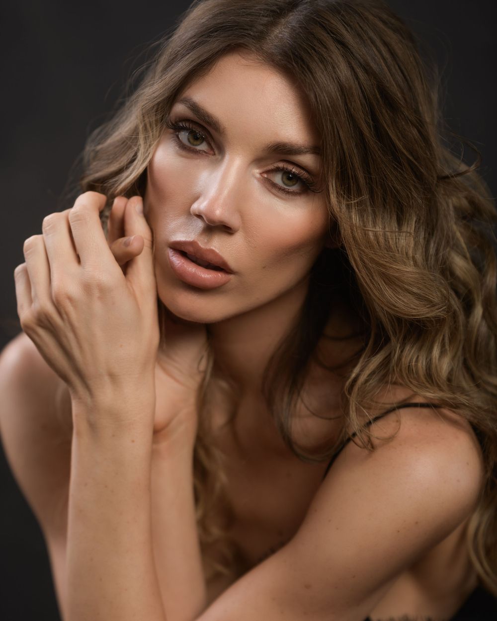 A beautiful woman with wavy brunette hair poses gracefully, hands near her face, against a dark background.