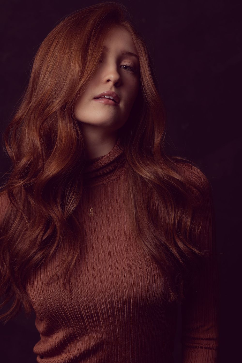 Young woman with long wavy auburn hair wearing a ribbed turtleneck sweater posed against a dark background.