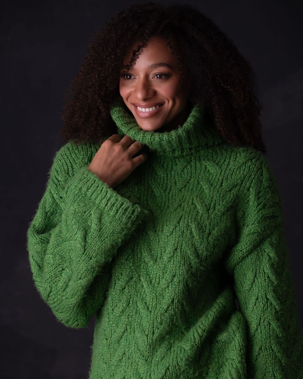 Beautiful portrait against dark background featuring someone in a chunky knit green turtleneck sweater with natural curly hair.