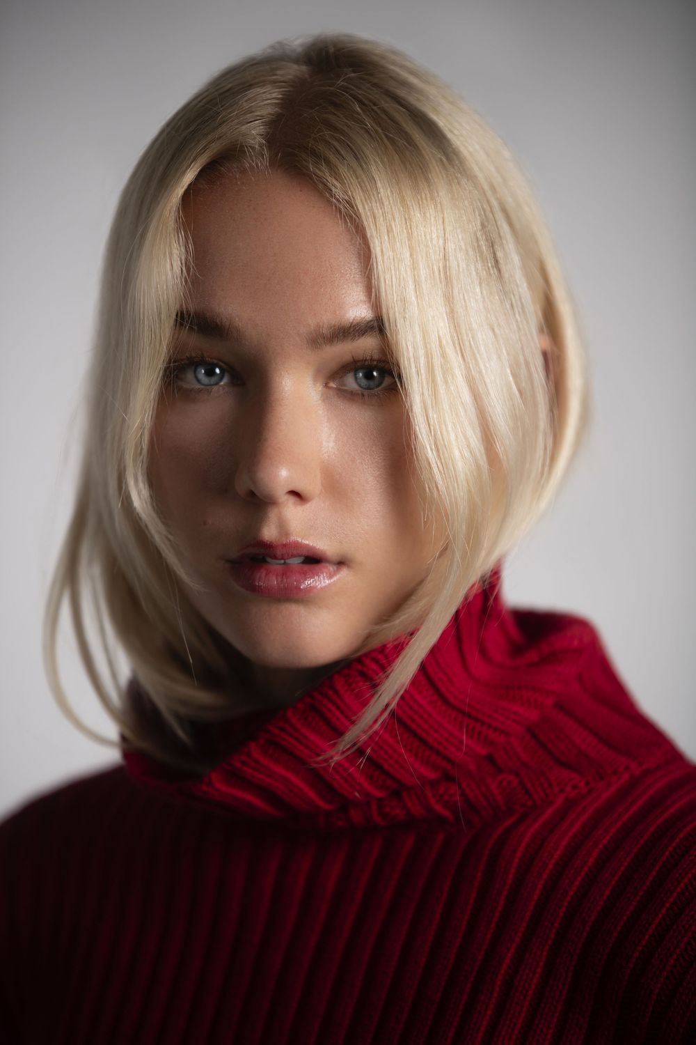 Young blonde woman with blue eyes wearing a red turtleneck sweater poses against a light gray background.
