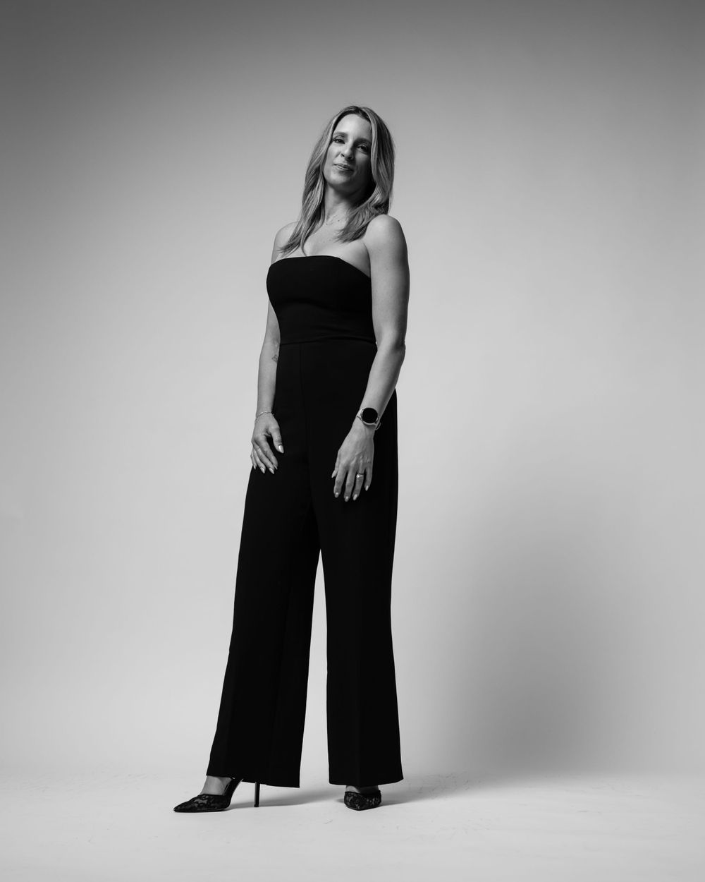 Black and white studio portrait of someone in a sleek black jumpsuit striking a sophisticated pose against plain backdrop.