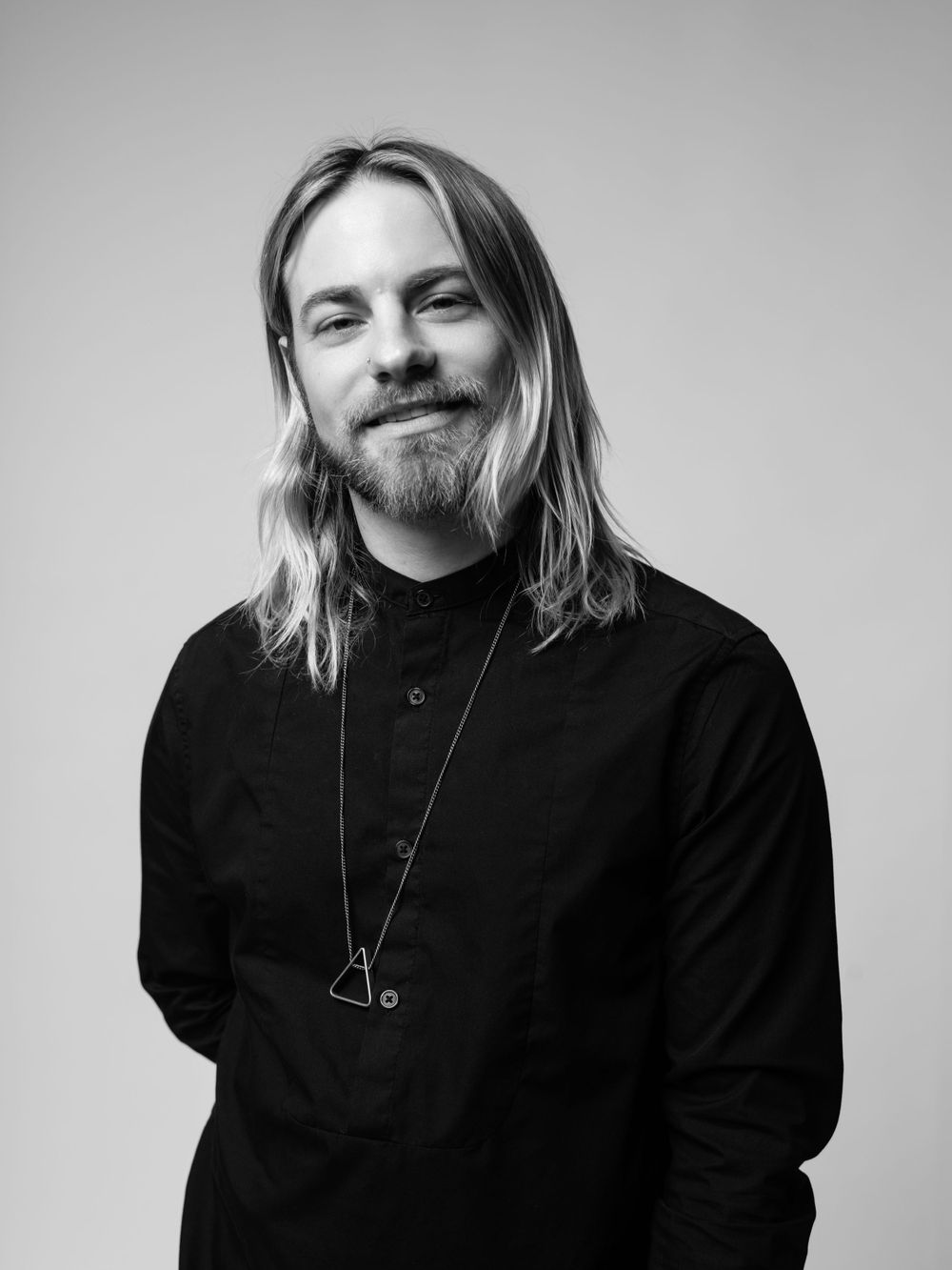 Black and white portrait of a person with long blonde hair wearing a black sweater and pendant necklace against a light background.