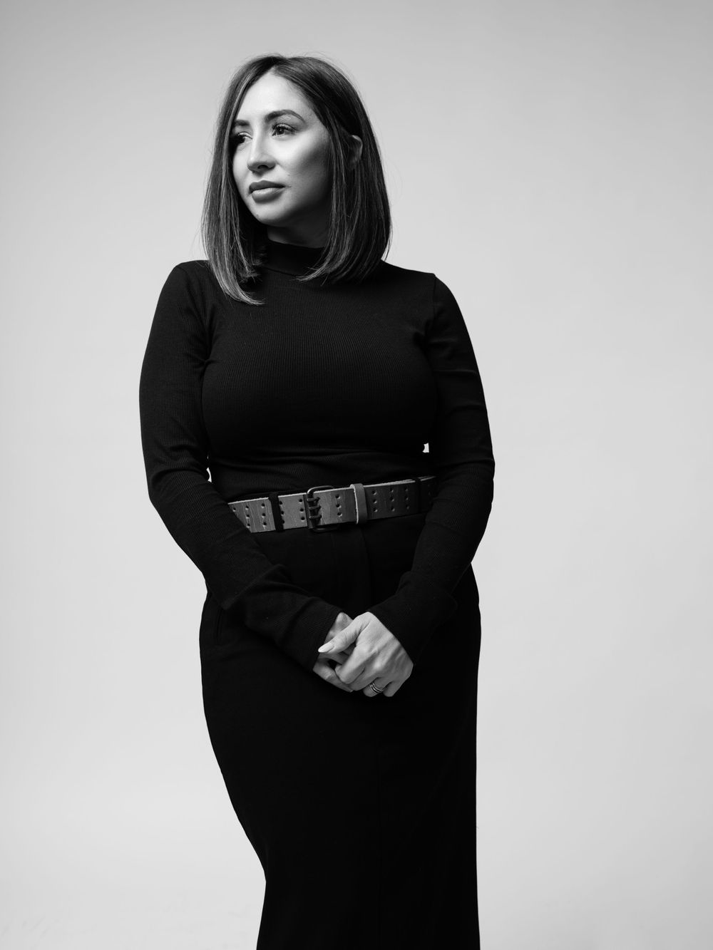 Black and white portrait photo of someone wearing a sleek black dress with a wide belt against a light background.