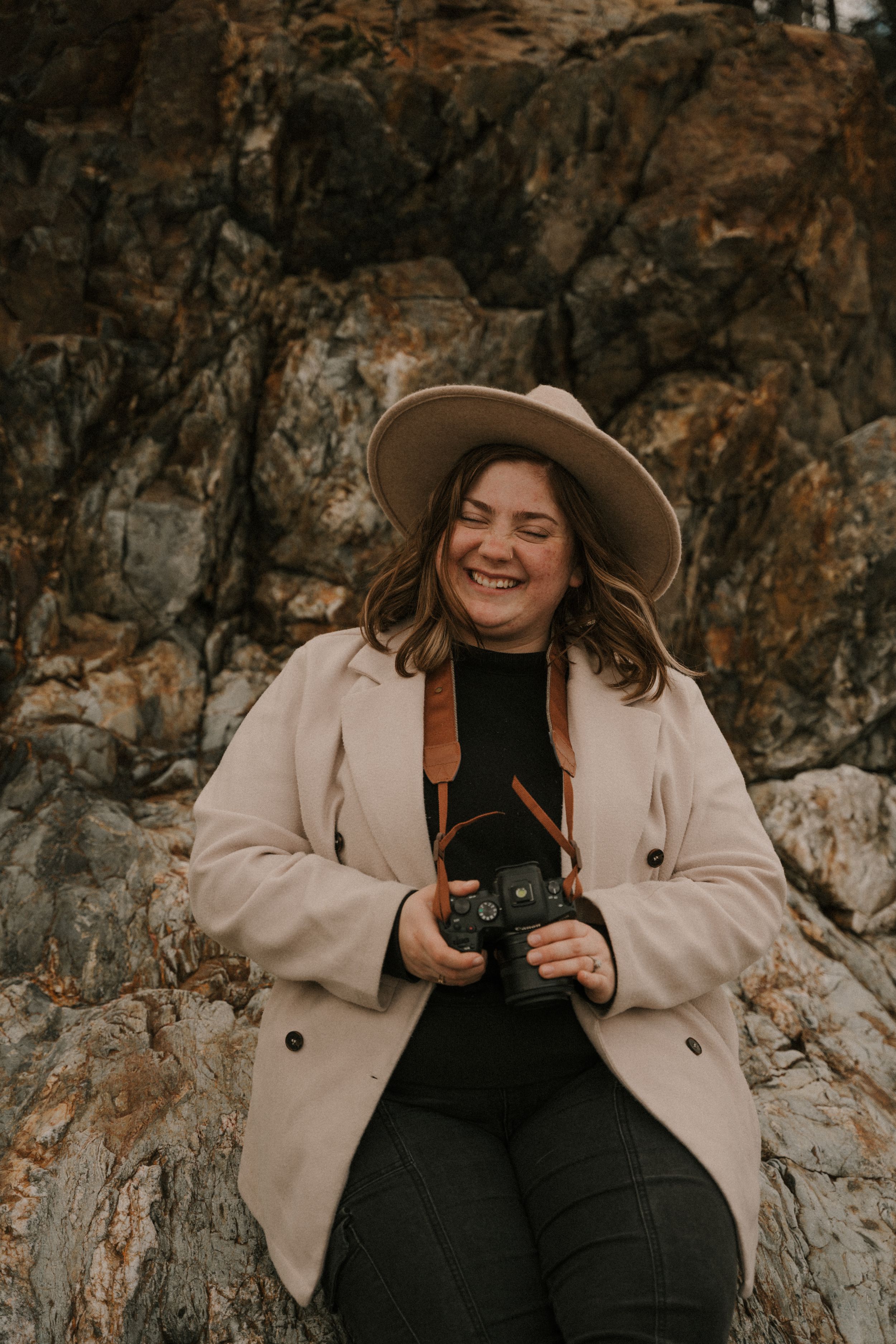 Person in beige coat and brown hat sitting against rocky cliff wall.