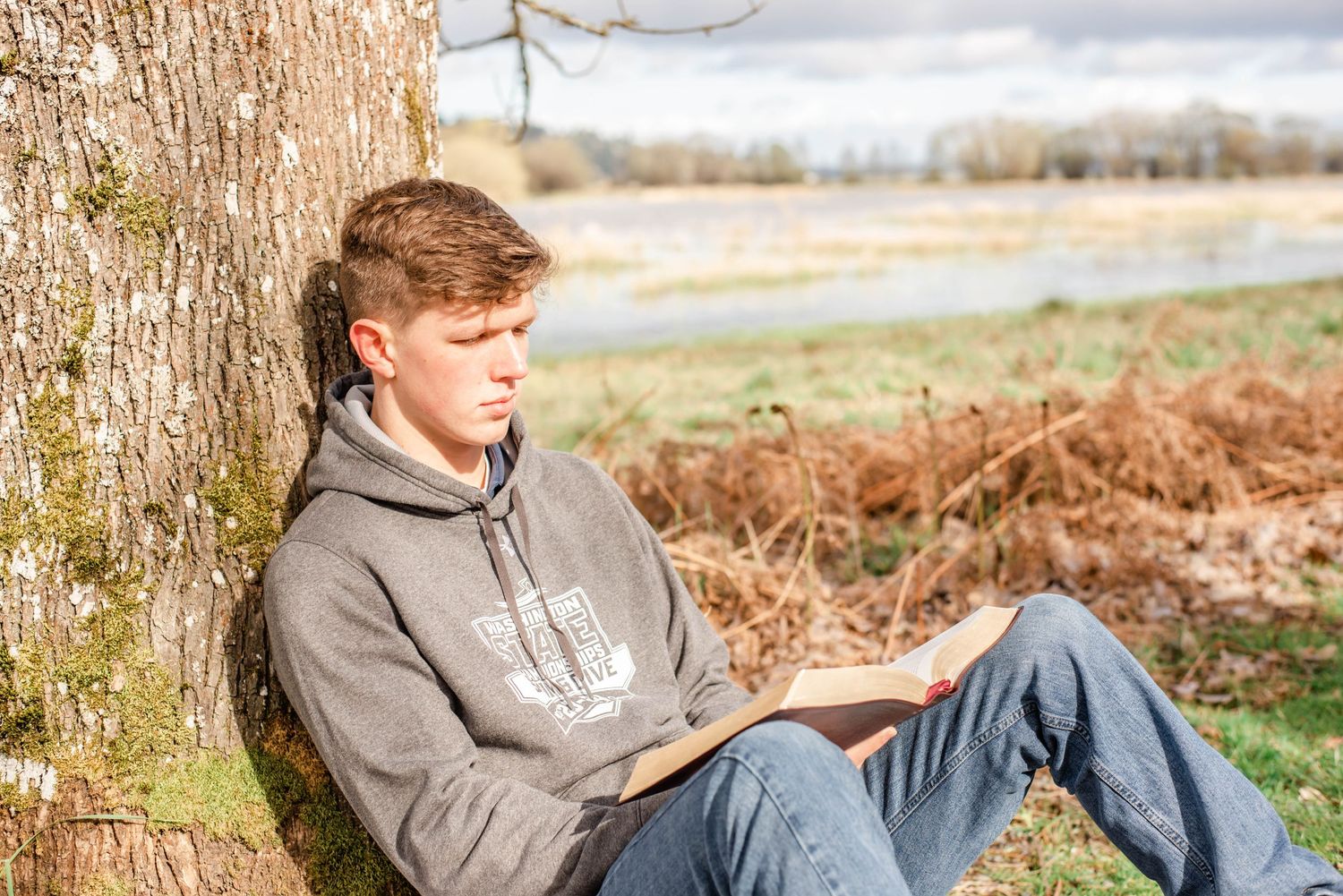 Person in grey hoodie reclining against tree trunk in outdoor setting.