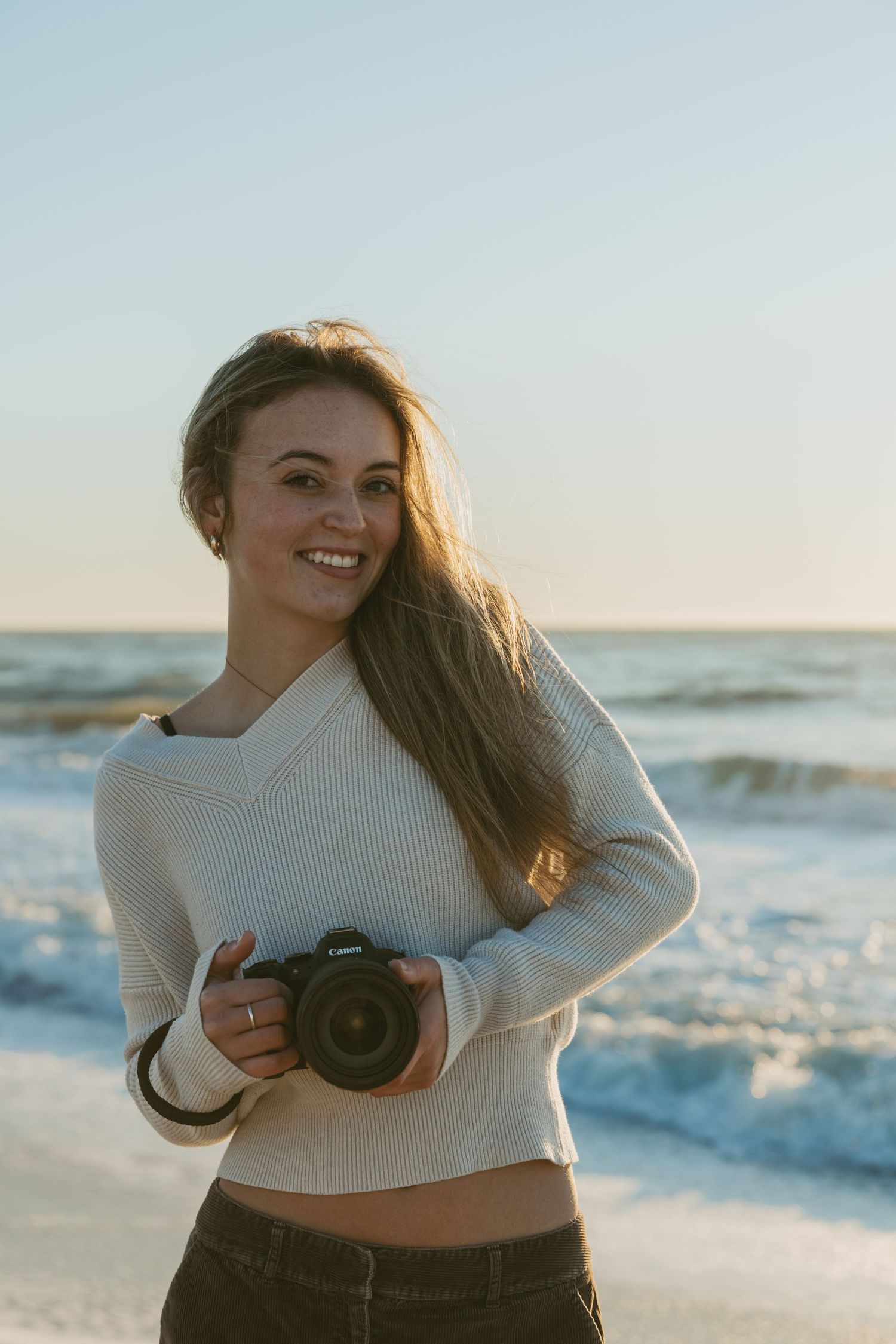 A person in a cream sweater poses with a camera at the beach during golden hour.