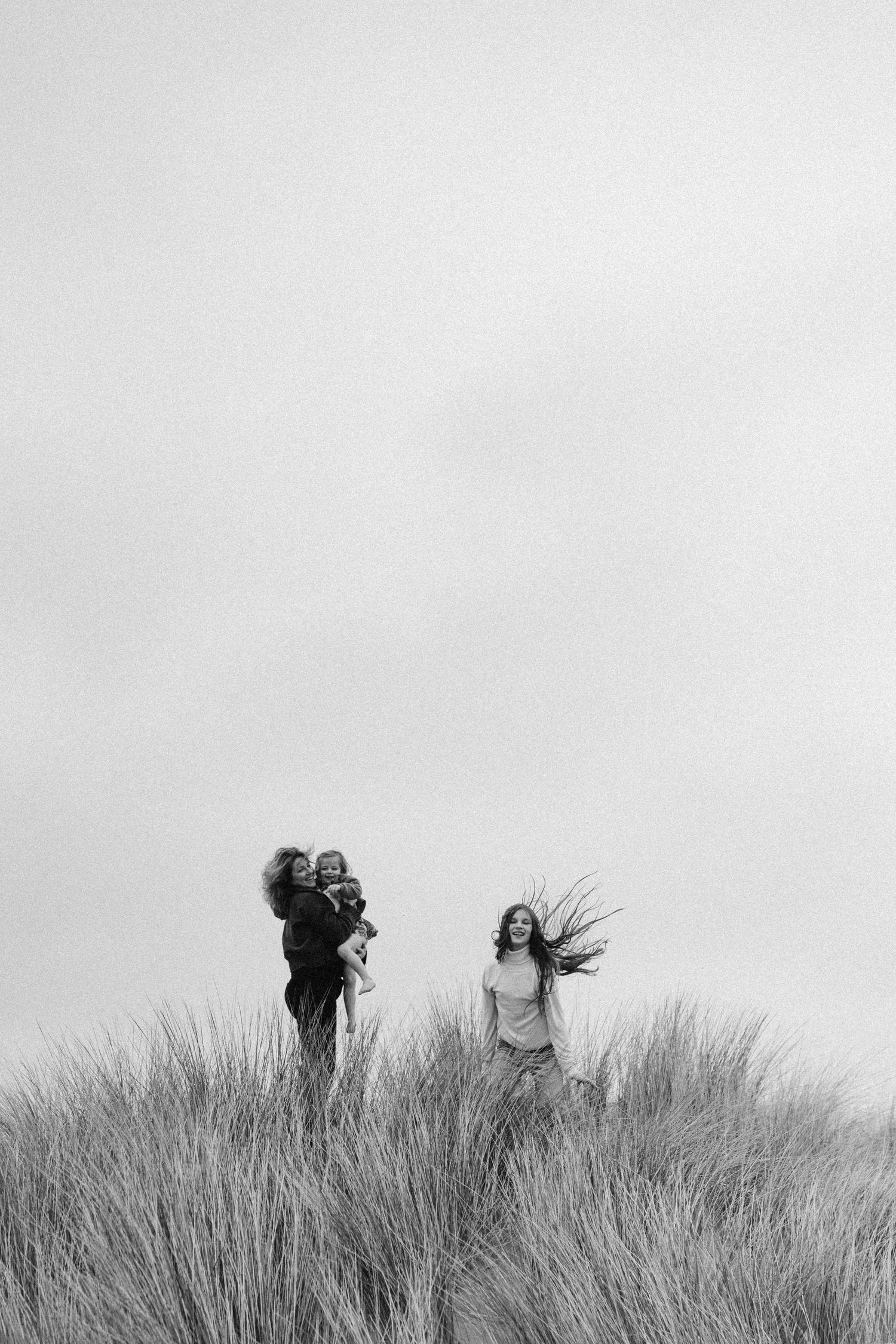 A mother standing on a sand dune, laughing and letting their hair to be blown away by the wind