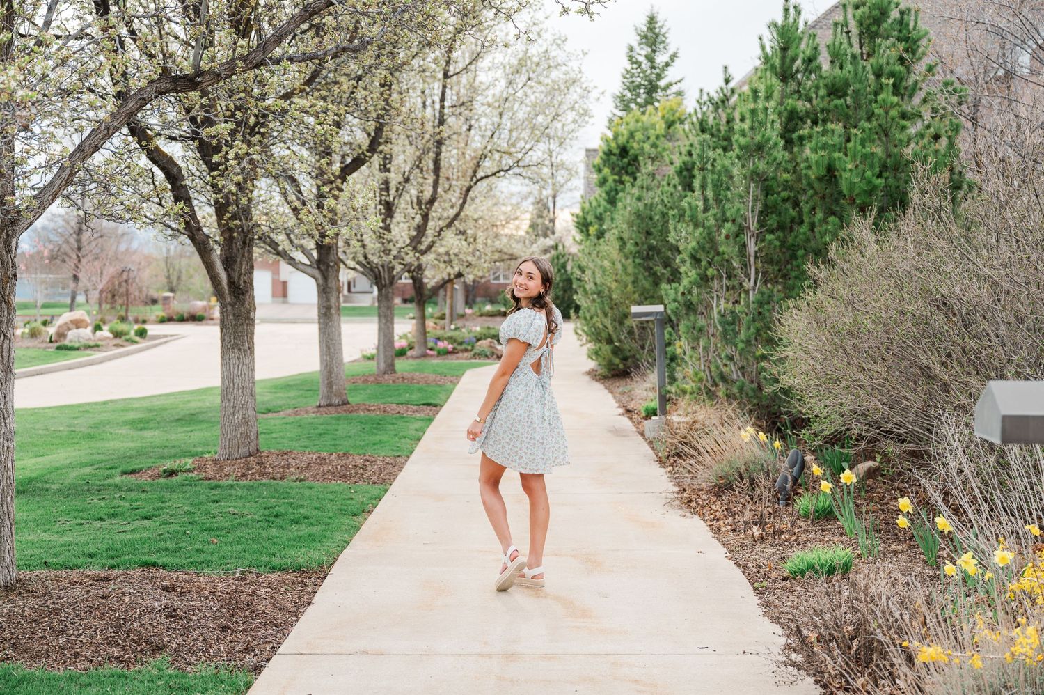 Person in white dress walks along sunlit pathway surrounded by trees and landscaping.