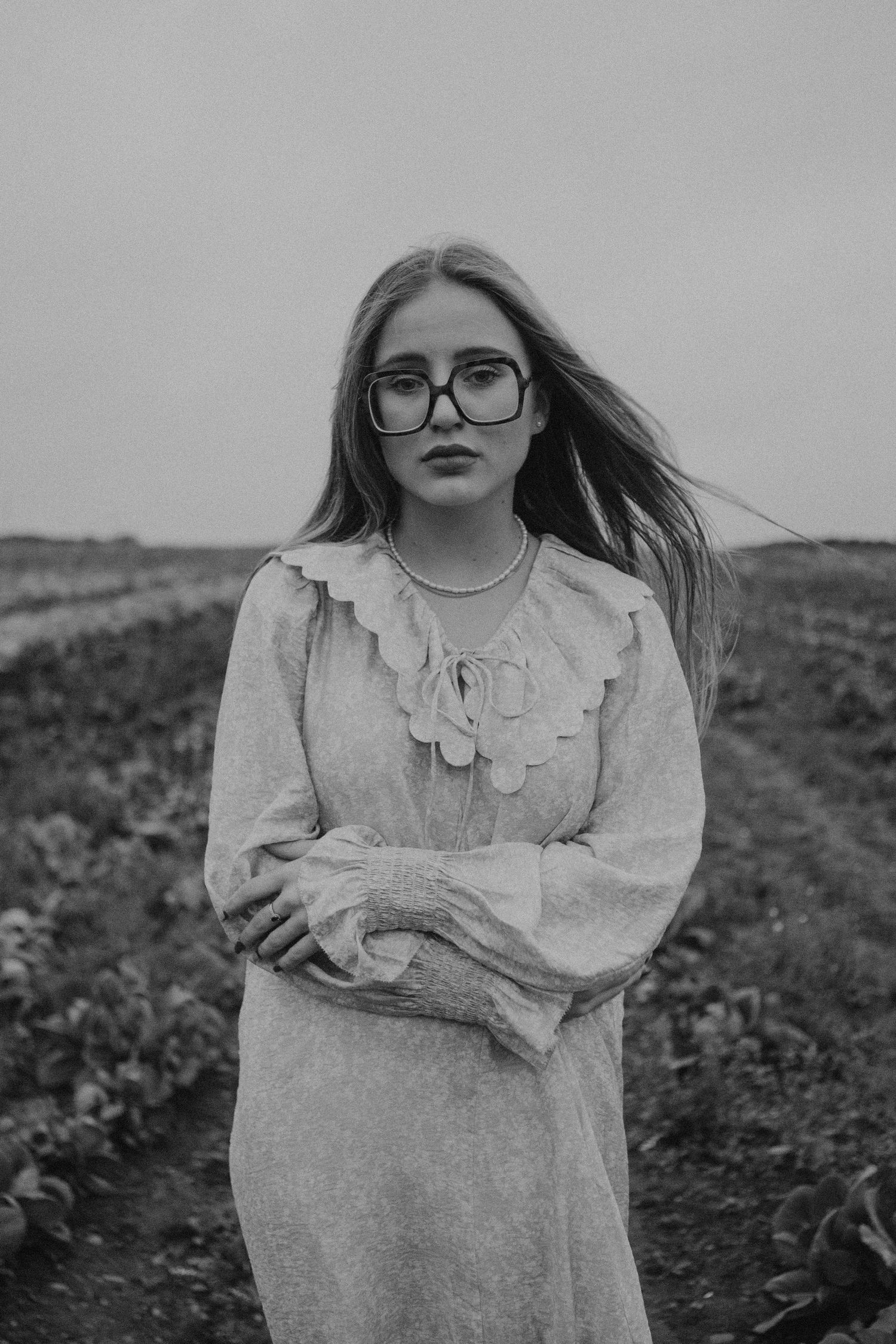 Person in vintage-style white dress with ruffles stands in field wearing round glasses.