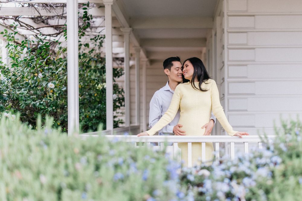 Couple standing on white columned porch with blue flowers blooming in garden below.