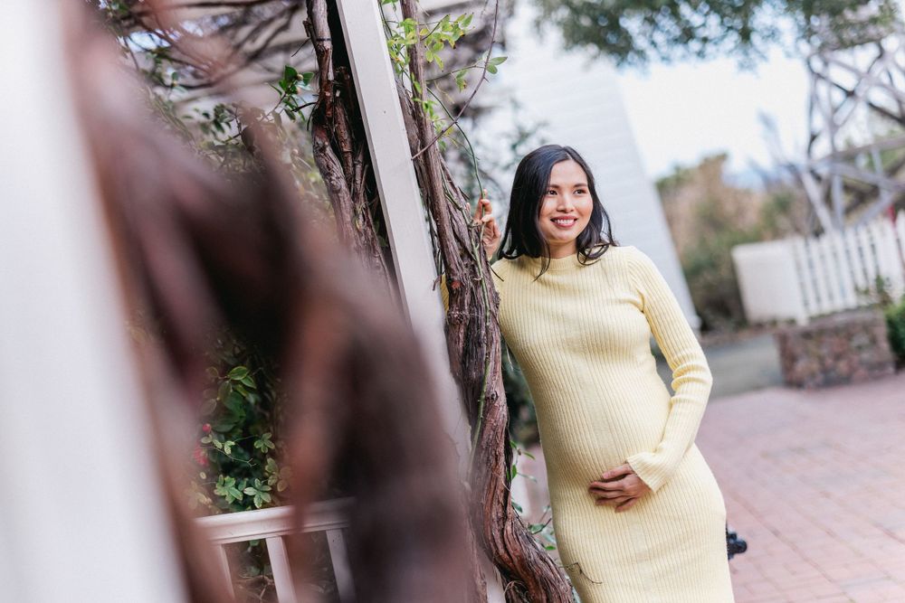 Expectant mother in yellow sweater dress poses against rustic wall during maternity shoot.