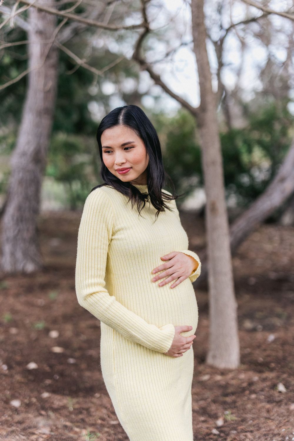 Woman in pale yellow ribbed sweater dress standing outdoors among trees.