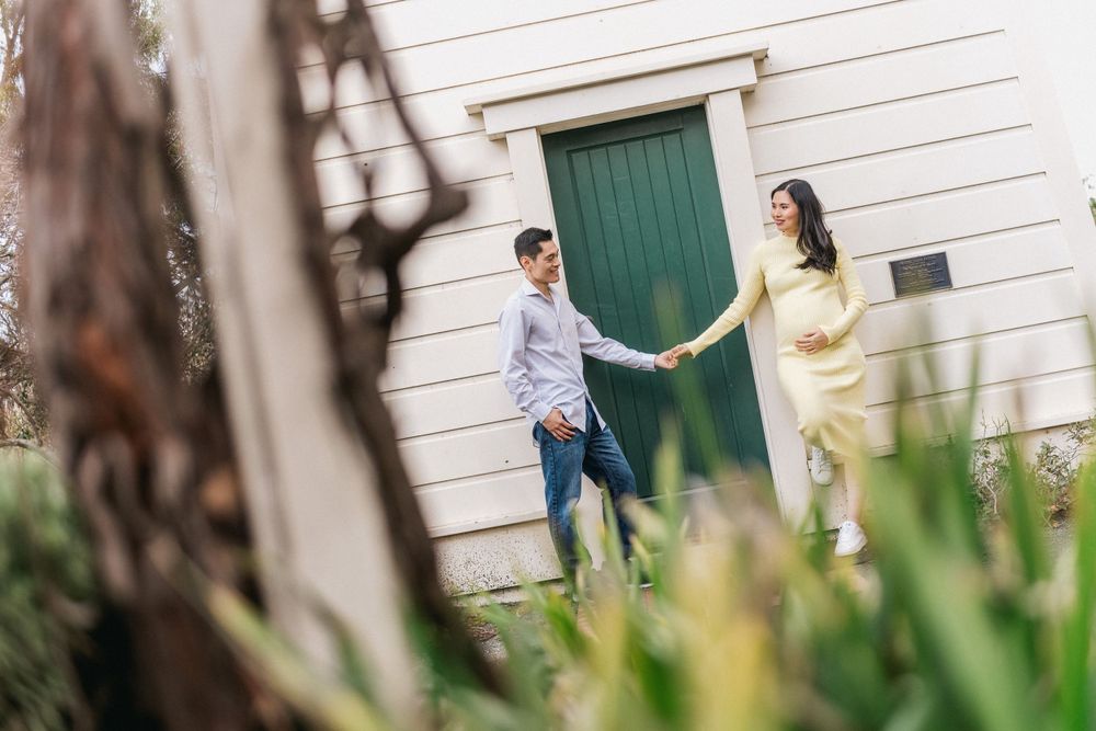 Couple holding hands by green door of white building framed by plants.