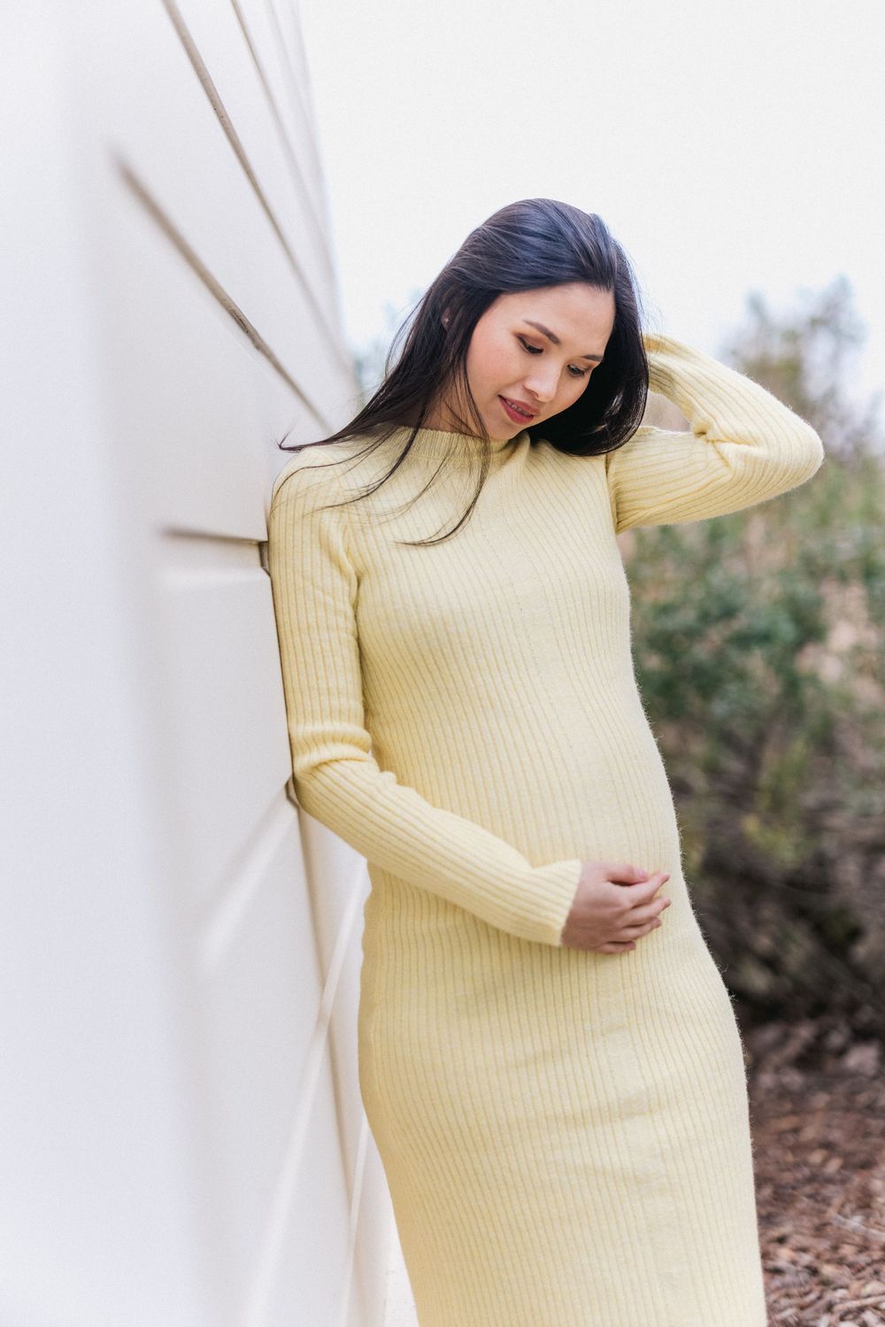 Woman in pale yellow sweater dress leaning against white wall outdoors.