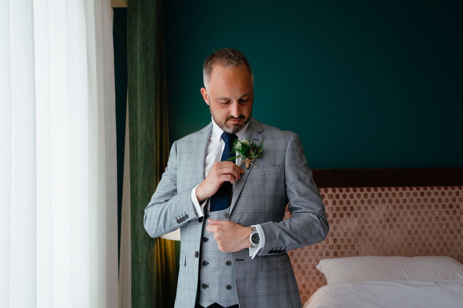 A person in a grey suit adjusts their boutonniere while getting ready.