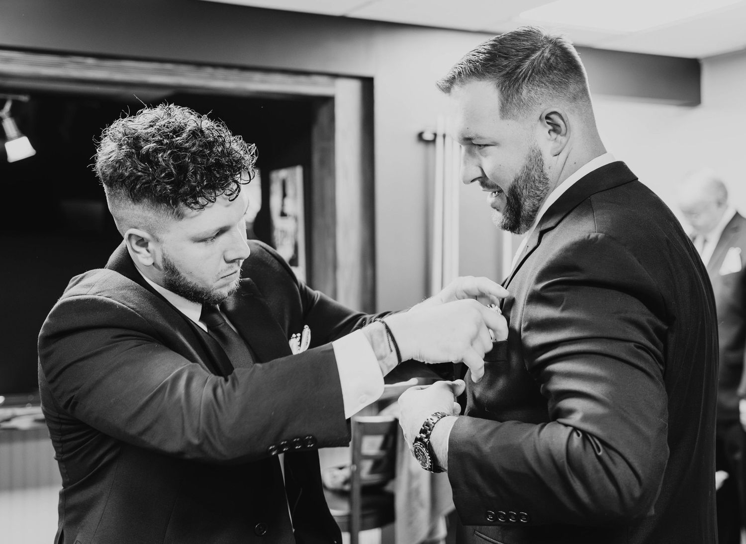 Two men in formal attire help each other with clothing adjustments in black and white photography.