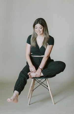 Casual portrait of someone sitting on a wooden stool wearing black loungewear and bare feet against neutral background.