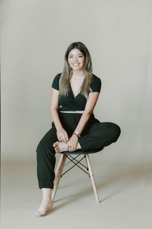 Relaxed lifestyle portrait showing person seated on wooden stool in black outfit against beige wall.
