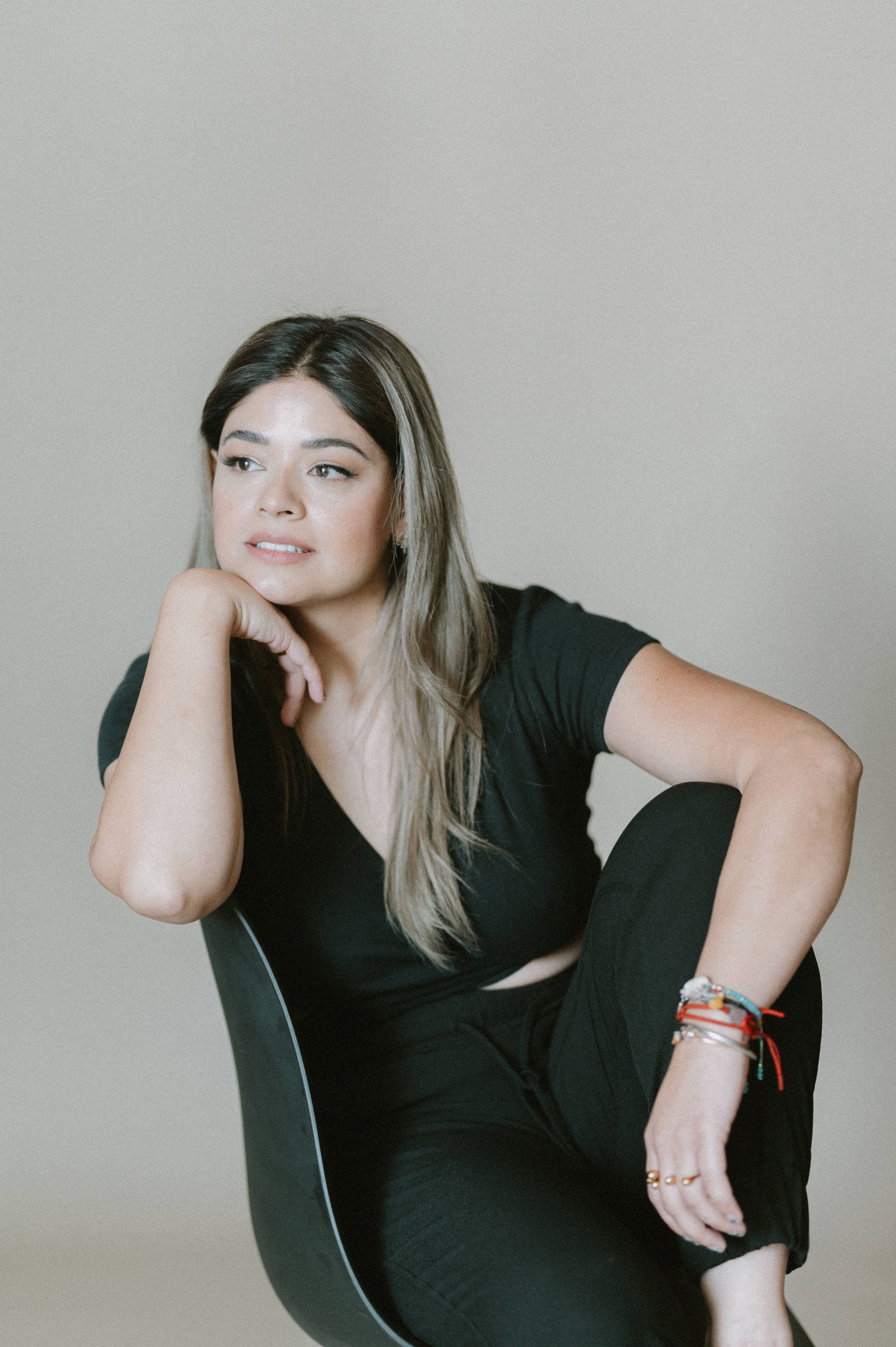 Model in black outfit poses on chair during minimalist studio photoshoot.