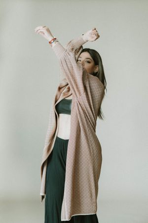 Energetic pose with arms raised wearing flowing beige cardigan and black athleisure against neutral background.