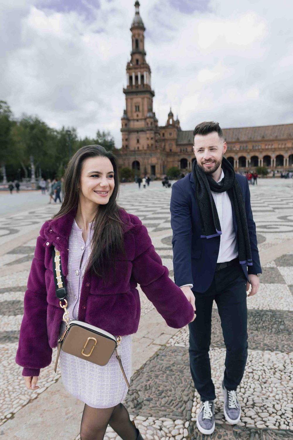 A couple holding hands at Plaza de España in Seville with architectural details in background.