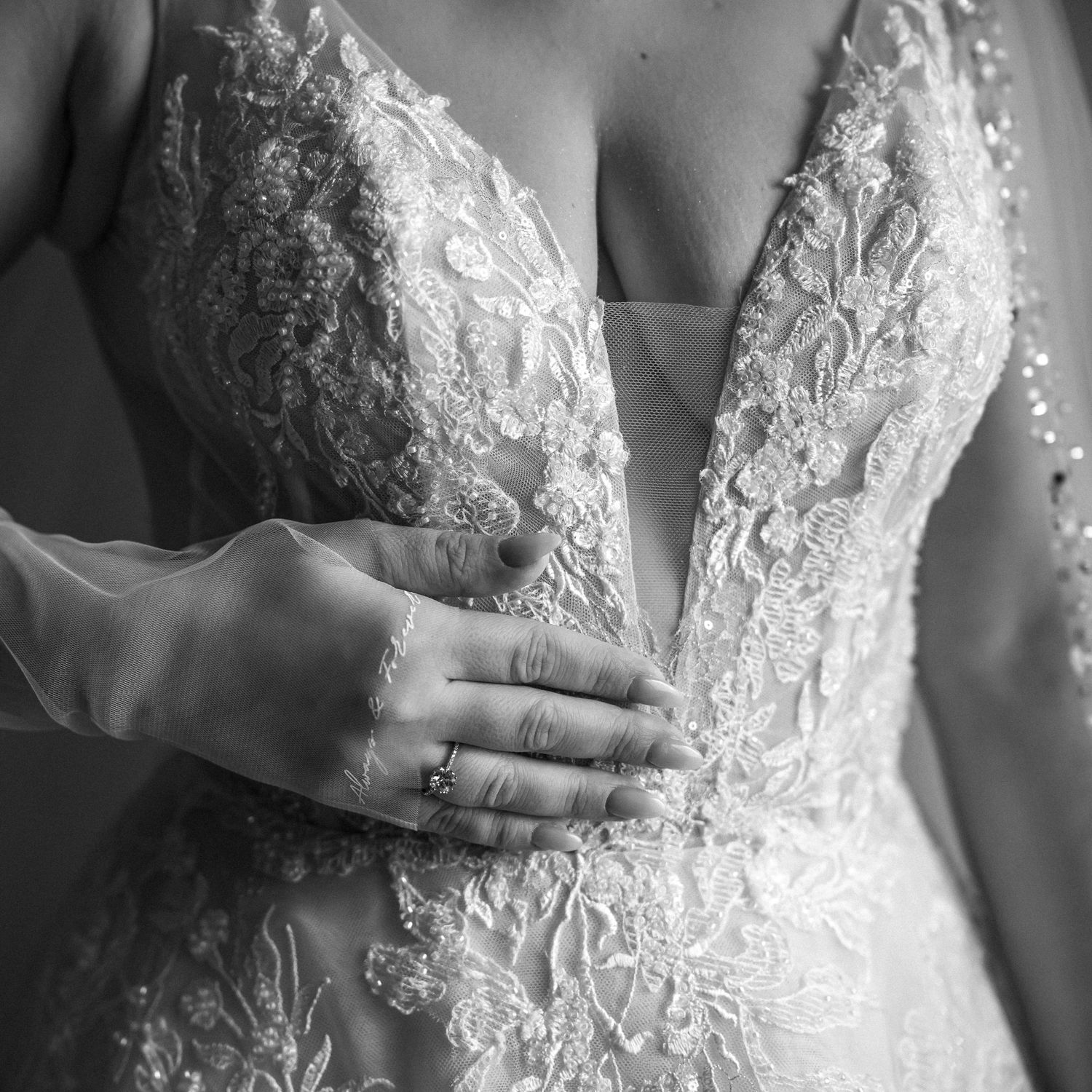 Close-up detail of intricate lace on a white wedding dress bodice with hand showing engagement ring.