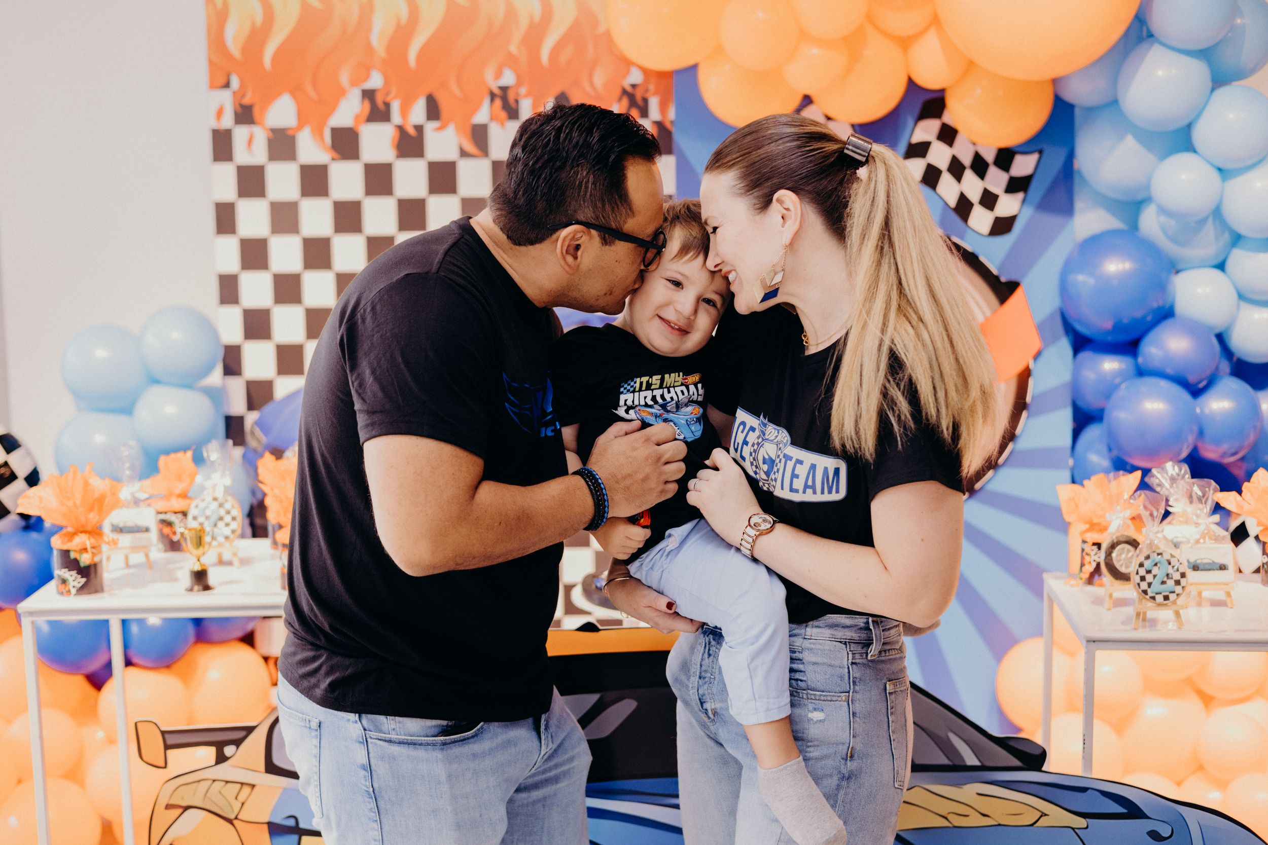 A family shares a joyful moment at a racing-themed birthday party with blue and orange balloon decorations.