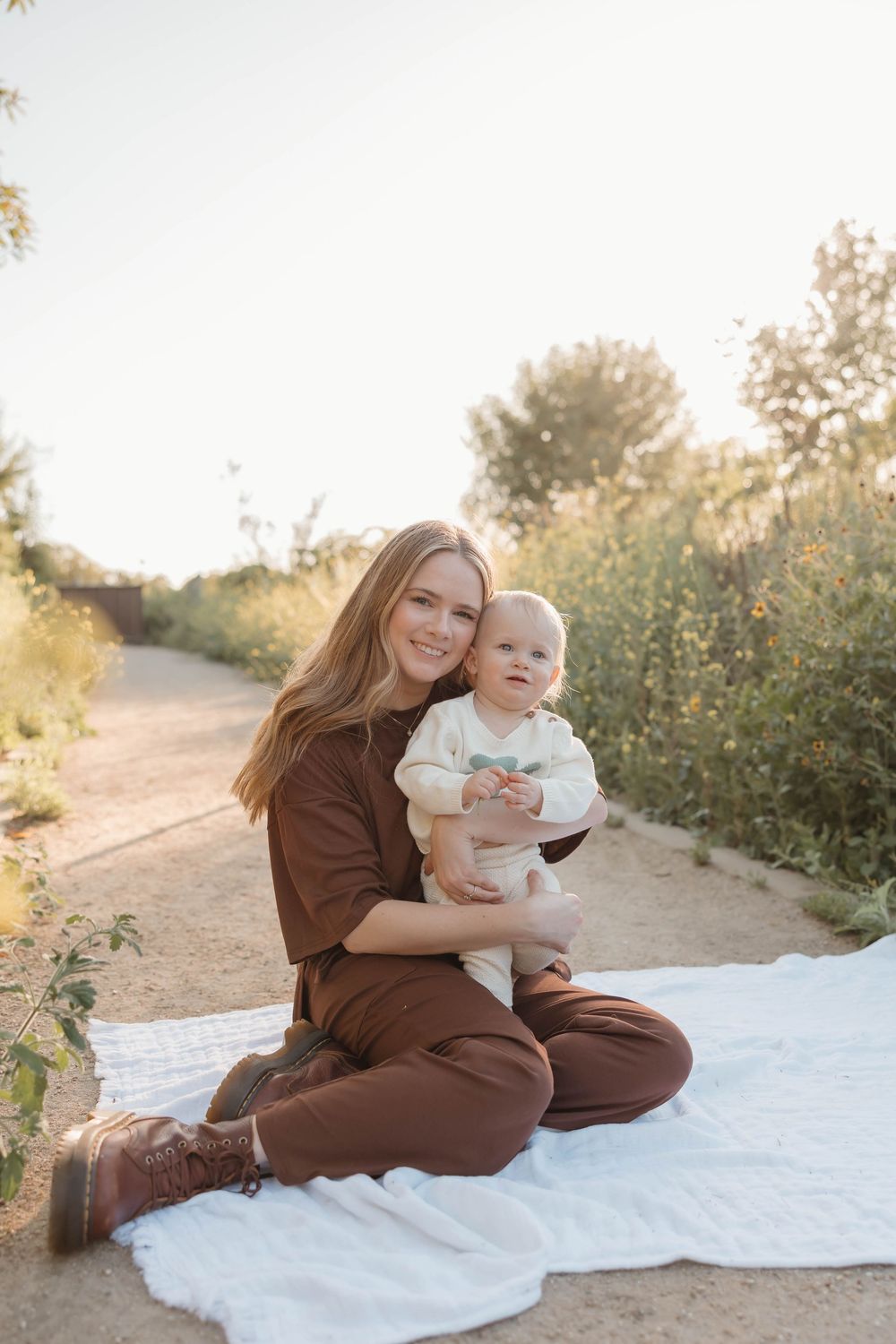 Young mother in brown outfit sits on white blanket outdoors, smiling while holding her baby in a sunny natural setting.