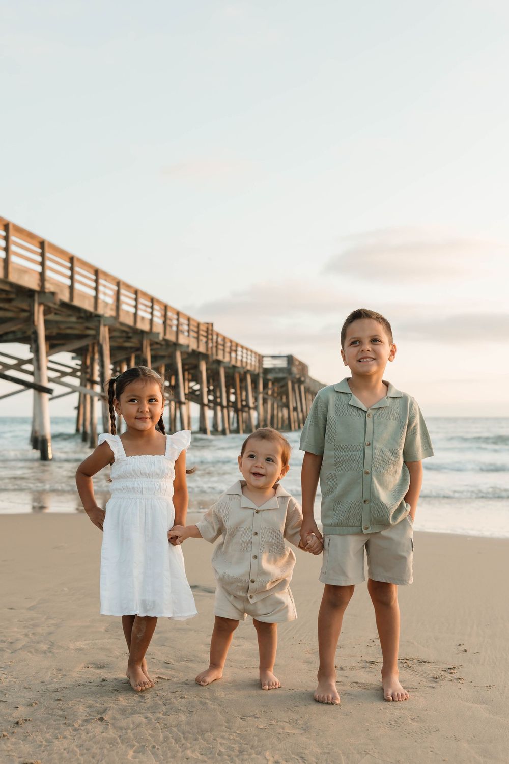 Children stand together on a sandy beach at sunset with a wooden pier in the background.