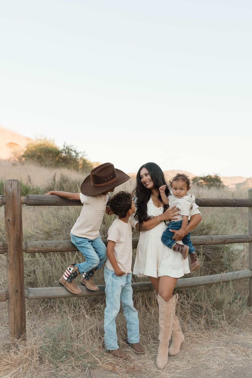 A woman in a white dress and cowboy boots holds a baby while a boy in a cowboy hat climbs a wooden fence outdoors.