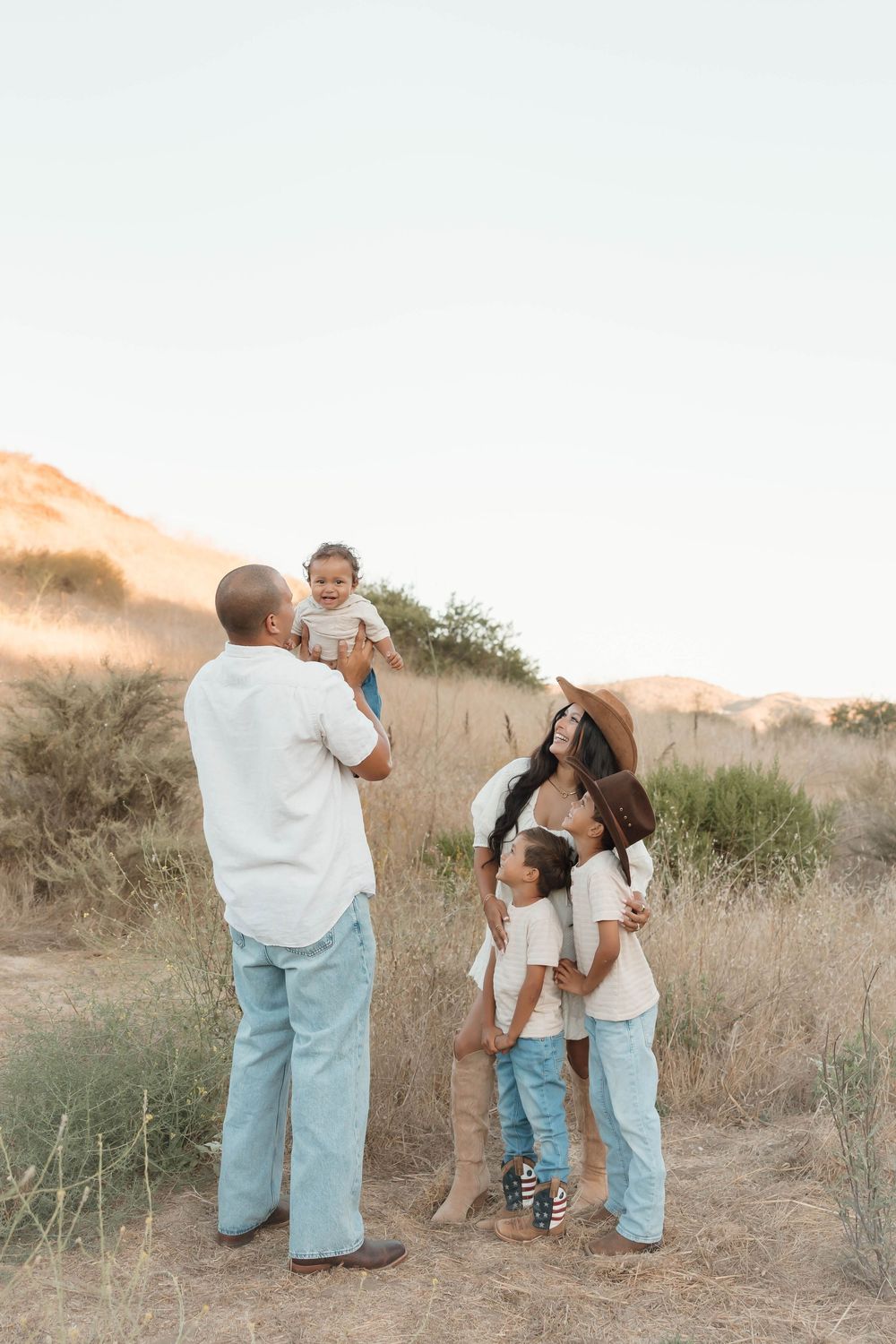 Family of five enjoying a golden hour outdoor photo session in a dry field with hills in the background.