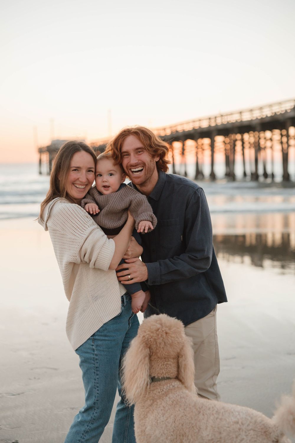 Family and their fluffy golden doodle dog share joyful moments together on a sunset beach near a pier.