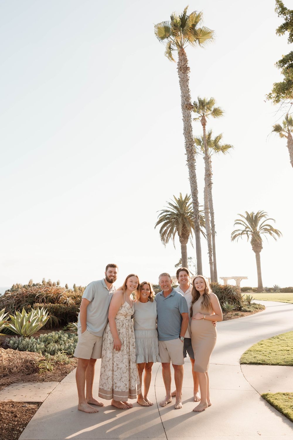Family of six posing on a sunny palm tree-lined path in a coastal setting, wearing neutral summer outfits.