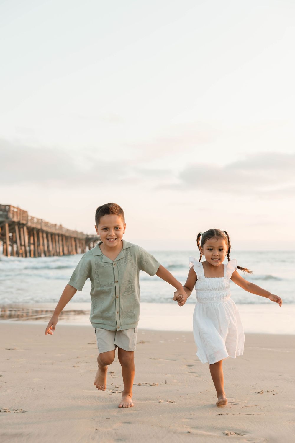 Two children in light colored outfits run and play together on a sandy beach at sunset.