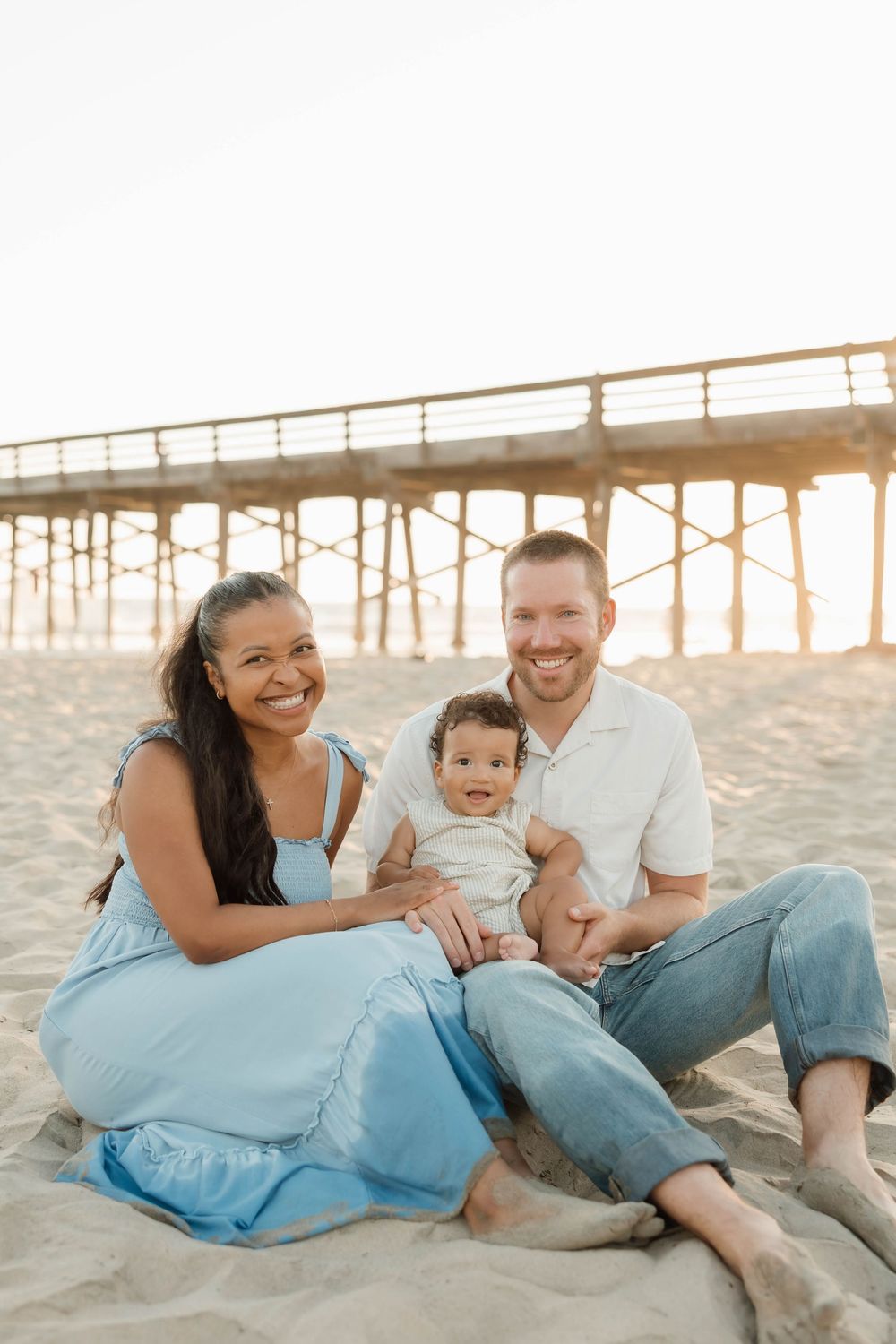 A happy interracial family of three sits on sandy beach near a pier, smiling warmly in golden sunset light.