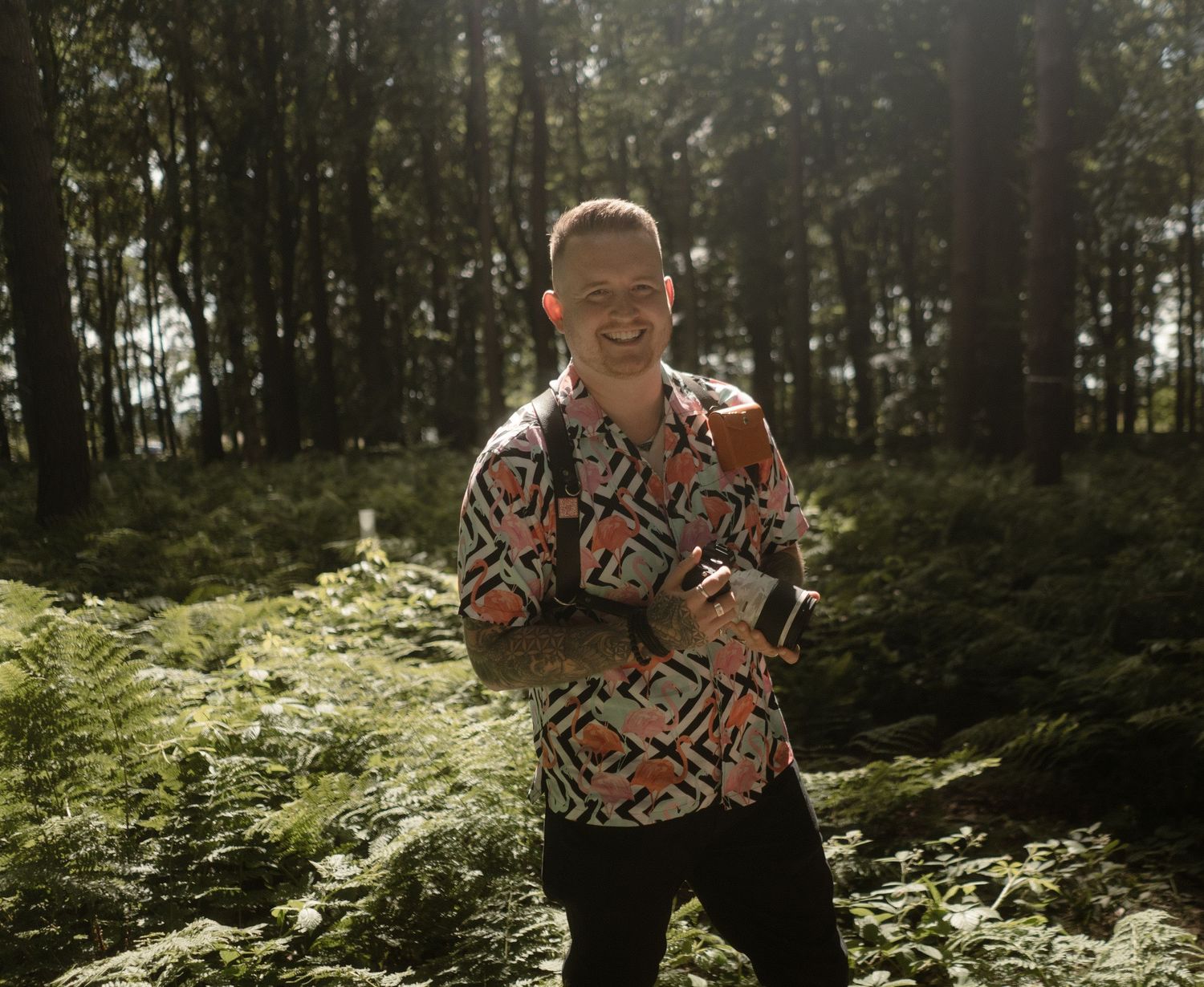 A photographer in a striking geometric pattern shirt poses with camera equipment in a lush forest.